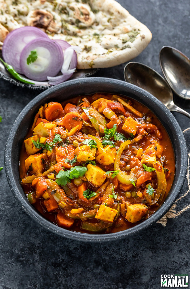 vegetable jalfrezi served in a black bowl with naan placed in the background along with some onion slices