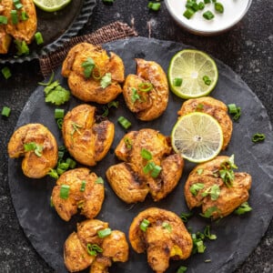 masala smashed potatoes served on a circular cheese board with lime wedges placed on the side. There's also a white color dip in a bowl in the background