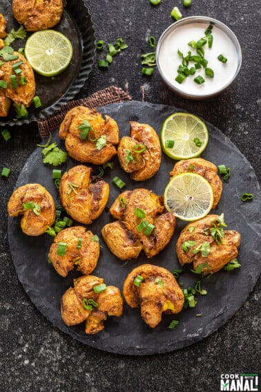 masala smashed potatoes served on a circular cheese board with lime wedges placed on the side. There's also a white color dip in a bowl in the background