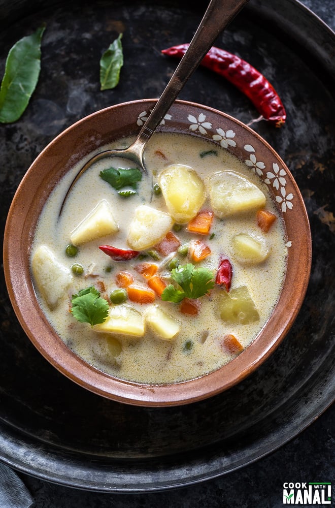 vegetable coconut stew in a serving bowl with a spoon, few curry leaves and dried chilies scattered in the background