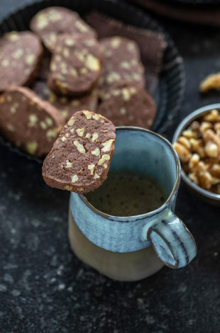 a chocolate walnut cookie placed on top of a coffee mug and a plate of cookies in the background