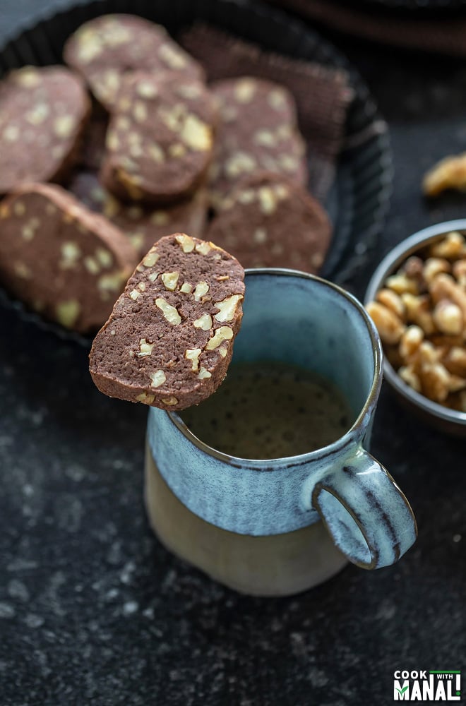 a chocolate walnut cookie placed on top of a coffee mug and a plate of cookies in the background