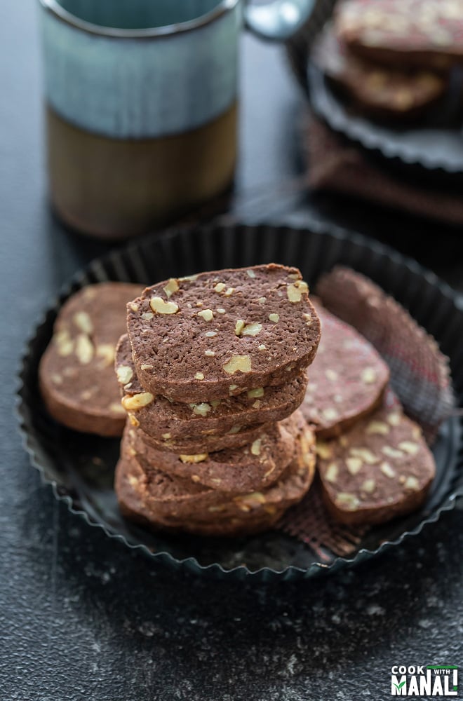 stack of chocolate walnut cookies placed in a rimmed black plate and a cup of coffee in the background