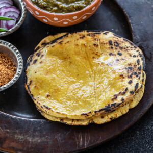 stack of makki ki roti on a plate with a bowl of saag and bowl of powdered jaggery and sliced onions on the sides