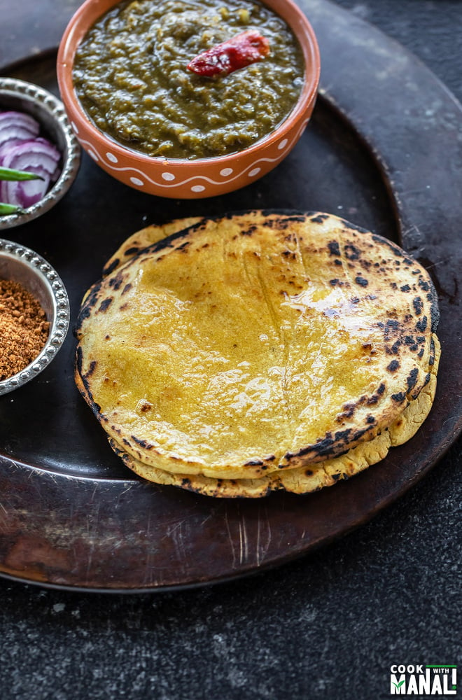 stack of makki ki roti on a plate with a bowl of saag and bowl of powdered jaggery and sliced onions on the sides