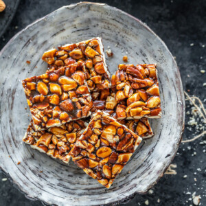 mixed nuts chikki in a white bowl and few nuts scattered on the sides and background