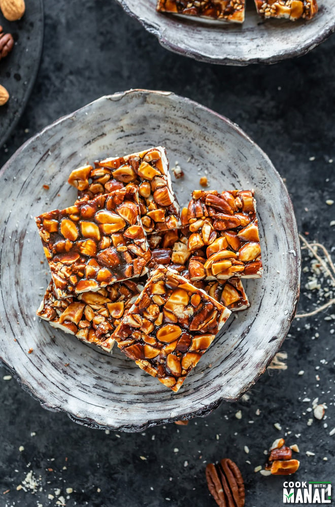 mixed nuts chikki in a white bowl and few nuts scattered on the sides and background