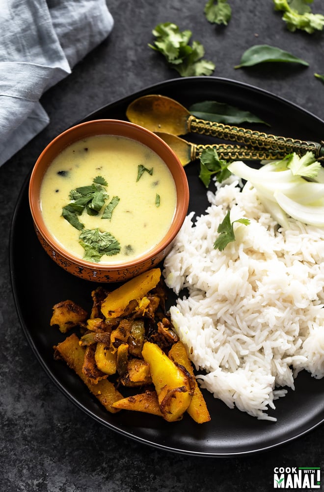 a black plate with a bowl of gujarati kadhi, boiled rice and spiced potatoes