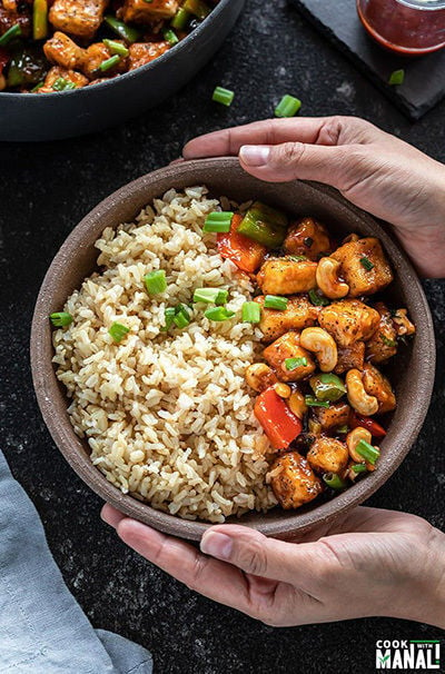 pair of hand holding a bowl of honey cashew tofu served with brown rice