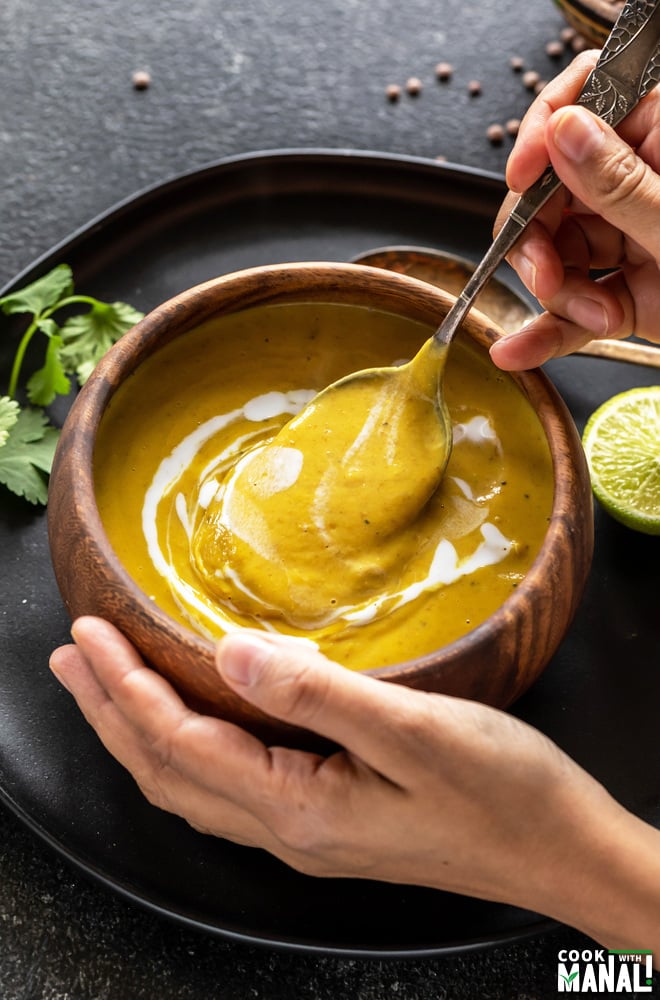 pair of hands holding a wooden bowl full of soup with one hand holding a spoon into the bowl