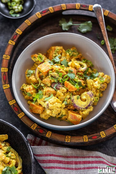 bowl of yellow color salad with cauliflower, sweet potato, onions and garnished with cilantro placed in a handcrafted serving tray