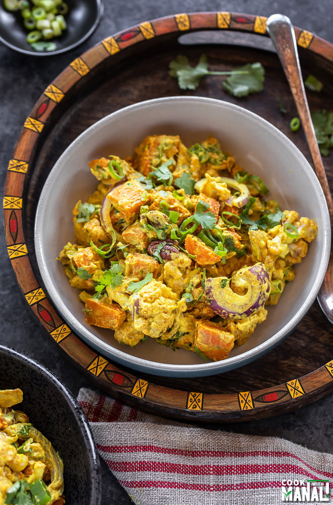 bowl of yellow color salad with cauliflower, sweet potato, onions and garnished with cilantro placed in a handcrafted serving tray