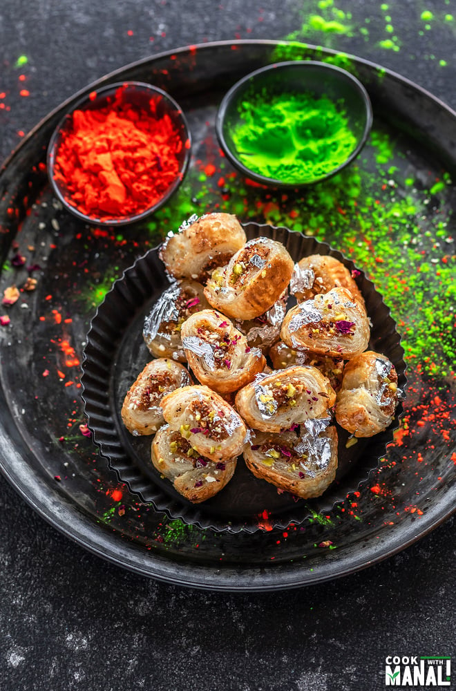 overhead shot of gujiya bites placed on a black rimmed plate with holi colors in the background