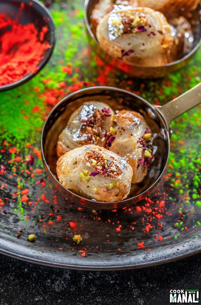 three pieces of gujiya bites (puff pastry dessert) placed in a copper bowl with another bowl in the background and colors on the side