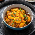 potato curry served in a bowl with a spoon and a plate of rice in the background