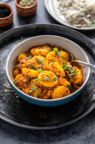 potato curry served in a bowl with a spoon and a plate of rice in the background