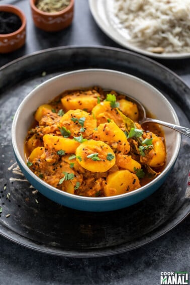 potato curry served in a bowl with a spoon and a plate of rice in the background