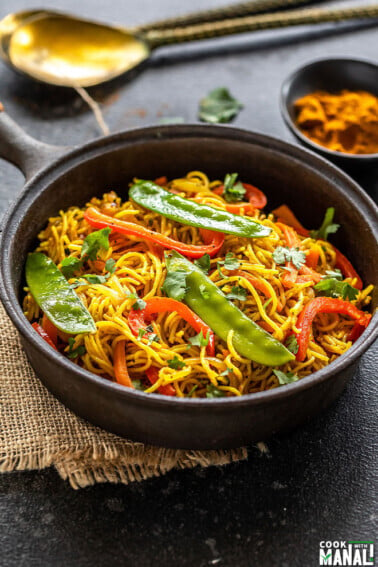 noodles with peppers, snow peas served in a black cast iron pan with a bowl of curry powder in the background along with two golden spoons