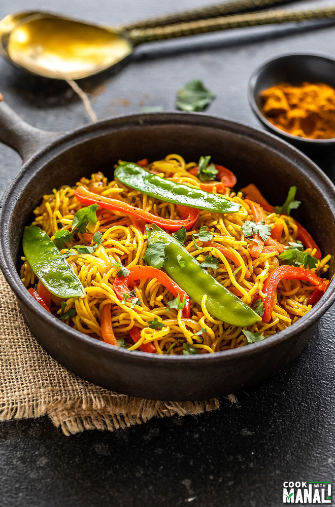 noodles with peppers, snow peas served in a black cast iron pan with a bowl of curry powder in the background along with two golden spoons