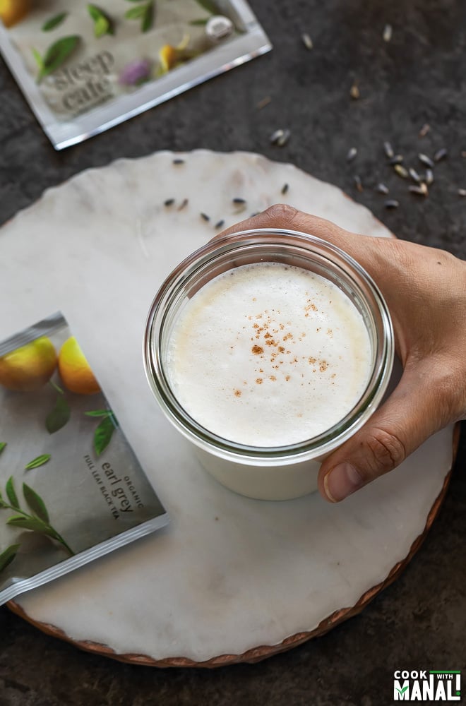 a hand holding a glass of latte with 2 sachet of tea bags lying on the side and background