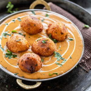 a copper serving ware with kofta (balls) placed on top of a orange color creamy looking gravy/curry and garnished with cilantro with a naan placed in the background