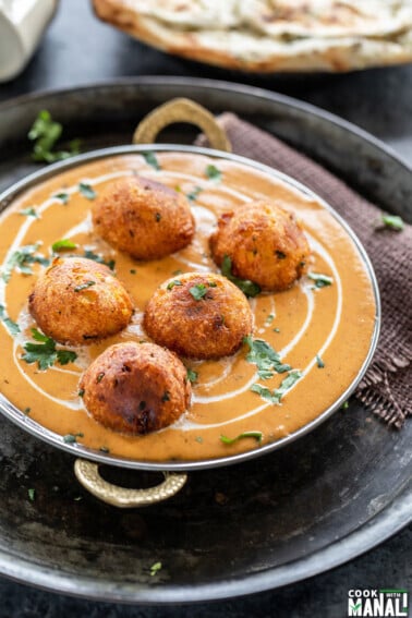 a copper serving ware with kofta (balls) placed on top of a orange color creamy looking gravy/curry and garnished with cilantro with a naan placed in the background