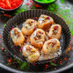 gujiya bites made from puff pastry arranged on a round plate and two bowls of holi color placed in the background