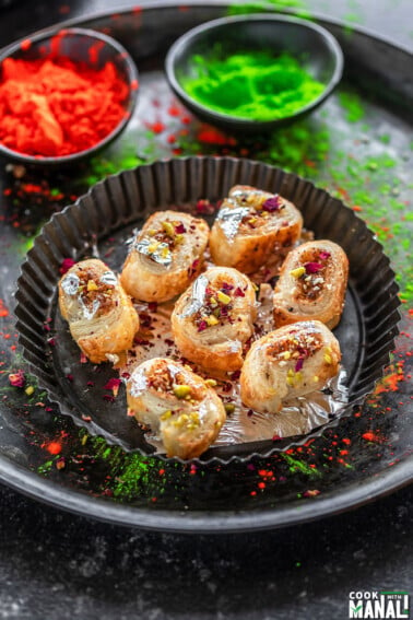 gujiya bites made from puff pastry arranged on a round plate and two bowls of holi color placed in the background