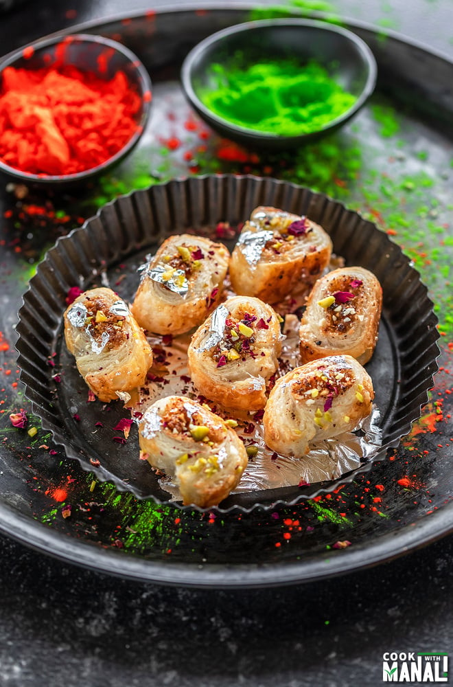 gujiya bites made from puff pastry arranged on a round plate and two bowls of holi color placed in the background