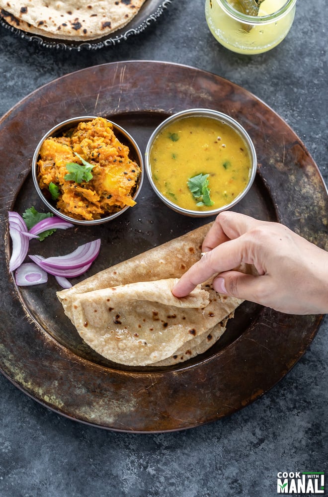 a hand breaking a piece of roti that is placed in a plate along with a bowl of dal and a potato curry
