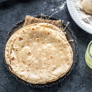 stack of roti placed in a silver antique looking plate with a rolling pin placed in the background and plate of wheat flour on the side