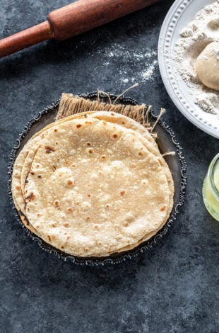 stack of roti placed in a silver antique looking plate with a rolling pin placed in the background and plate of wheat flour on the side