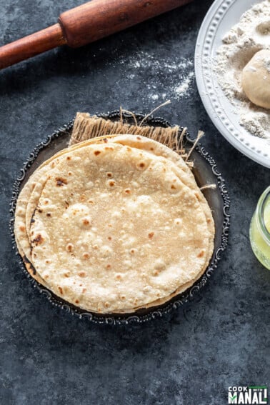 stack of roti placed in a silver antique looking plate with a rolling pin placed in the background and plate of wheat flour on the side