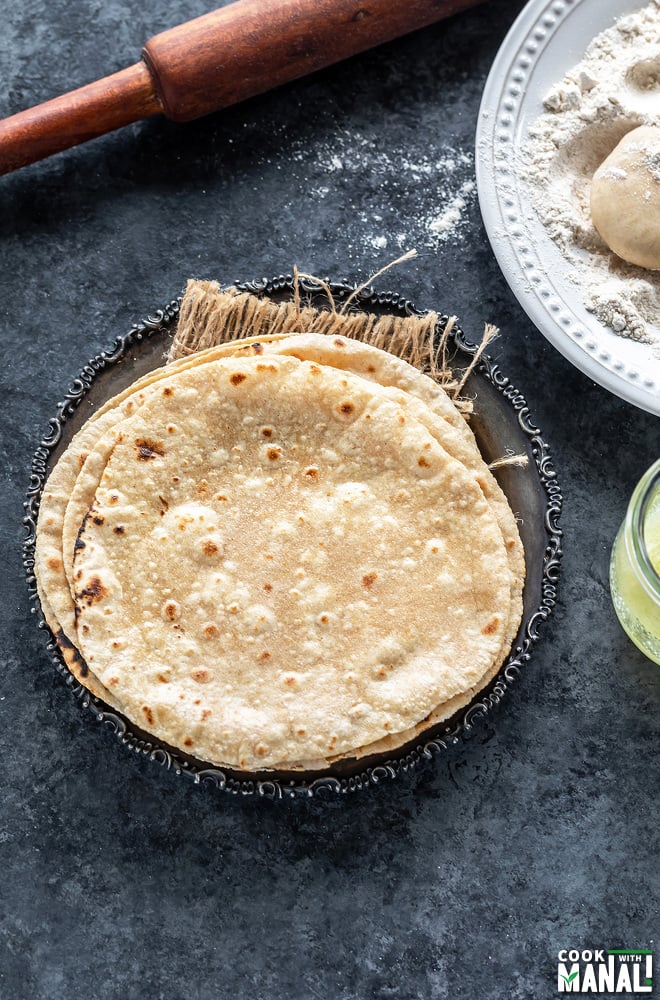 stack of roti placed in a silver antique looking plate with a rolling pin placed in the background and plate of wheat flour on the side