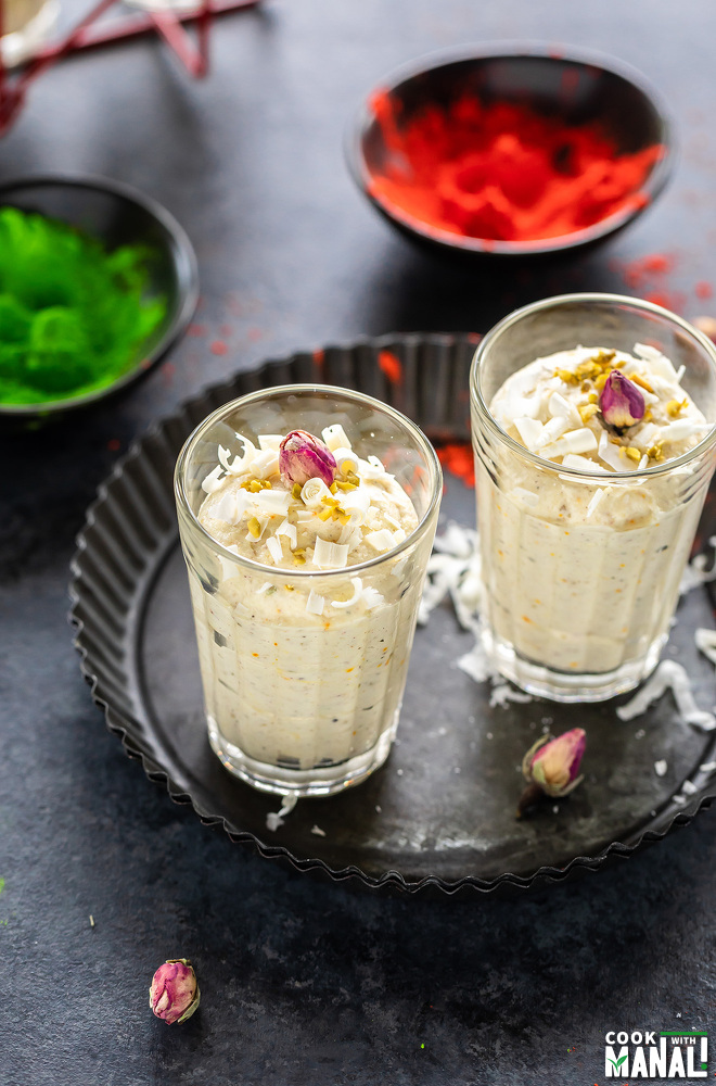 two glasses of thandai mousse placed in black rimmed plate with bowls of color in the background