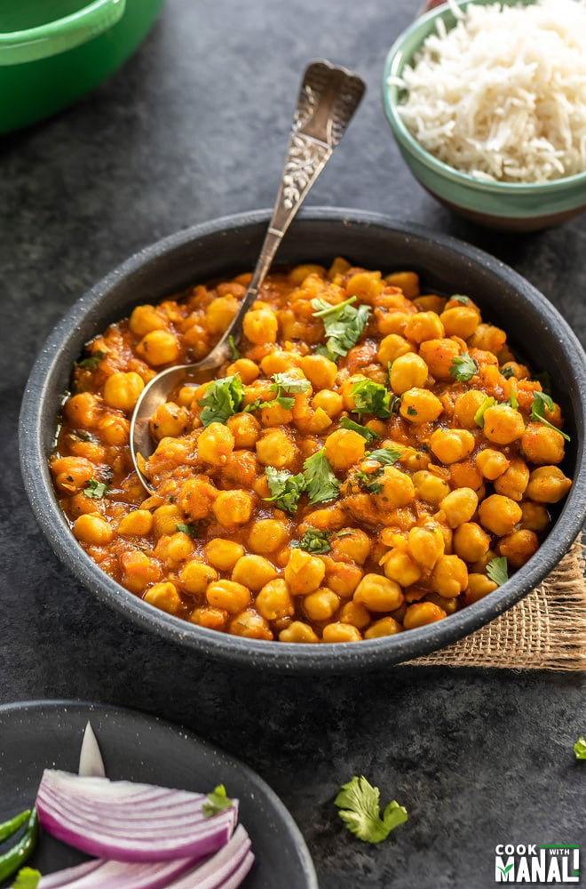 chana masala served in a round black bowl with a spoon, a small bowl of rice placed in the background and another plate with sliced onions on the side