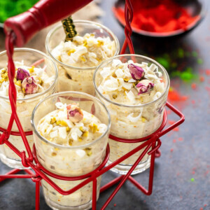 four glasses of white chocolate thandai mousse placed in a glass holder with bowls filled with red and green color in the background