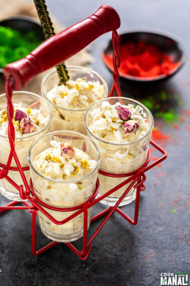 four glasses of white chocolate thandai mousse placed in a glass holder with bowls filled with red and green color in the background