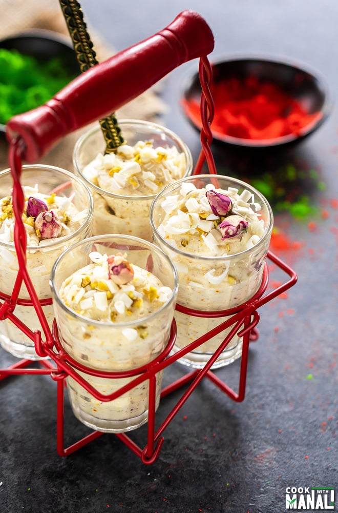 four glasses of white chocolate thandai mousse placed in a glass holder with bowls filled with red and green color in the background