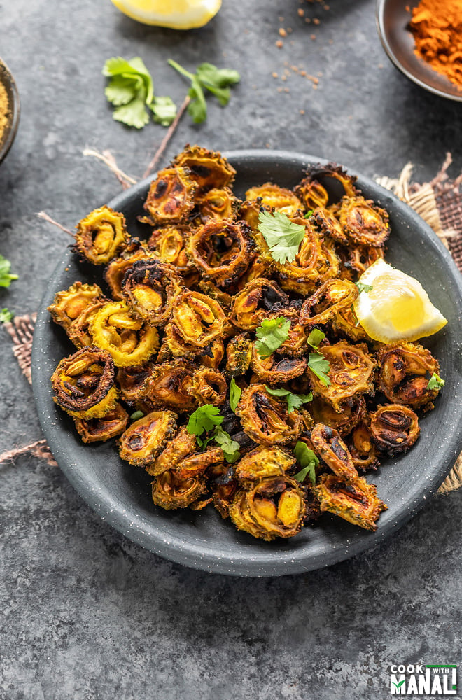 sliced karela with spices served in a black plate garnished with lemon wedge and cilantro