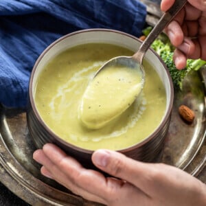 pair of hand holding a bowl of broccoli soup with few broccoli florets scattered in the background