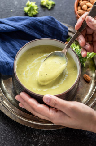 pair of hand holding a bowl of broccoli soup with few broccoli florets scattered in the background