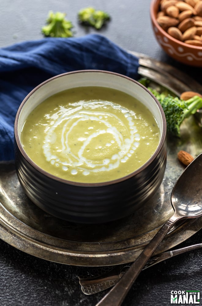 bowl of broccoli soup drizzled with milk with a spoon placed on the side and a bowl of almonds and few broccoli florets placed in the background