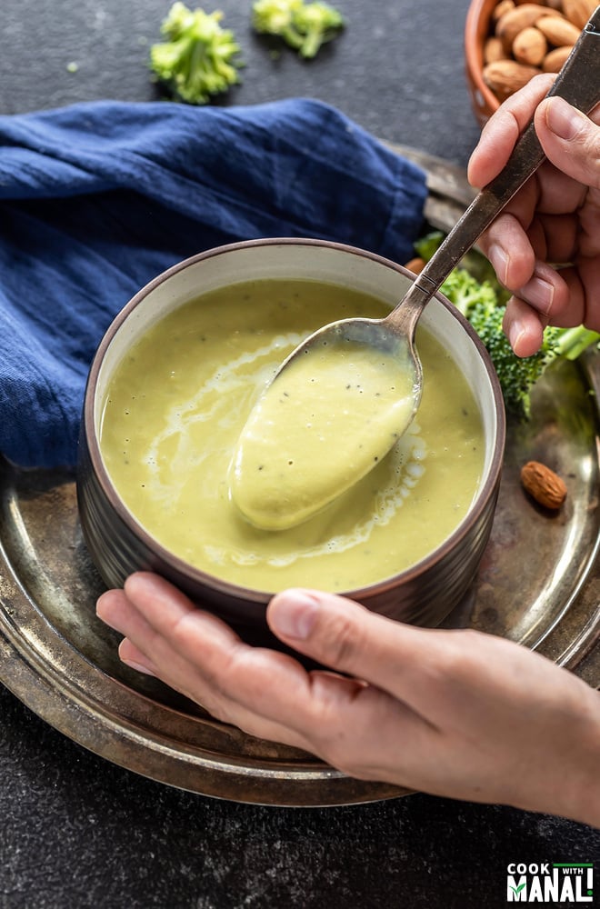 pair of hand holding a bowl of broccoli soup with few broccoli florets scattered in the background