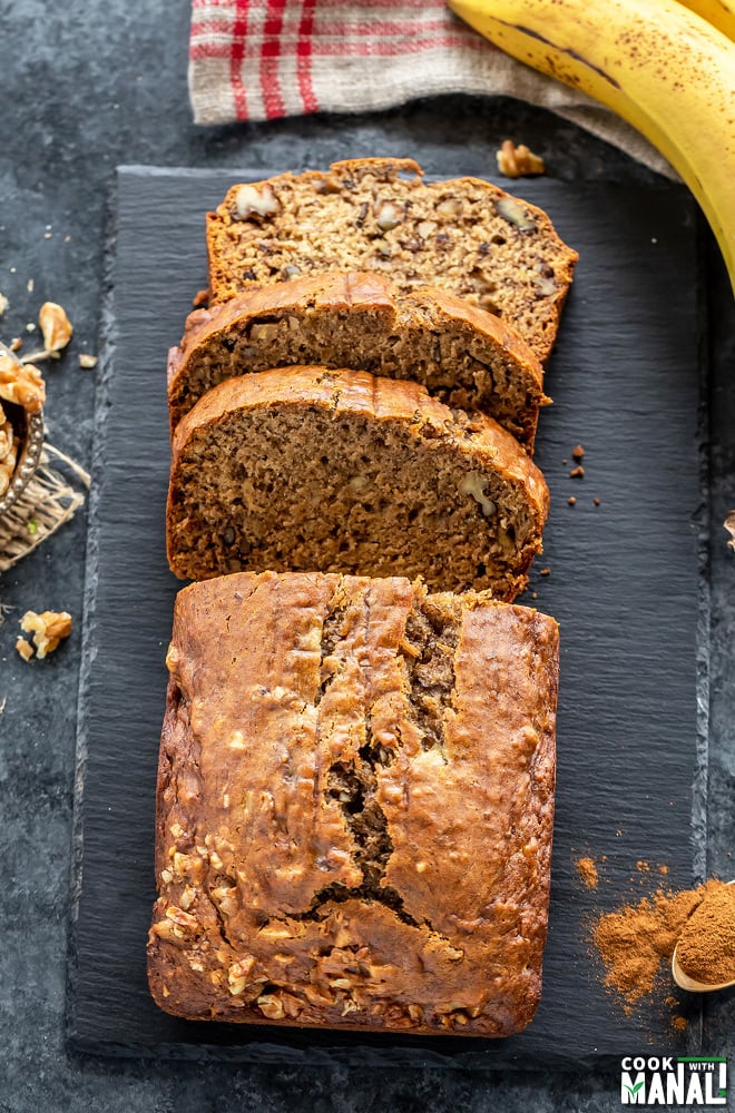 a banana bread with three cut slices placed on a black board with a banana placed on the side