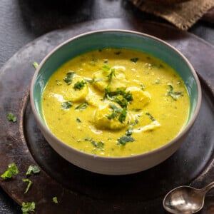 yellow color curry served in a bowl garnished with cilantro with two spoons placed on the side and two small bowls in the background