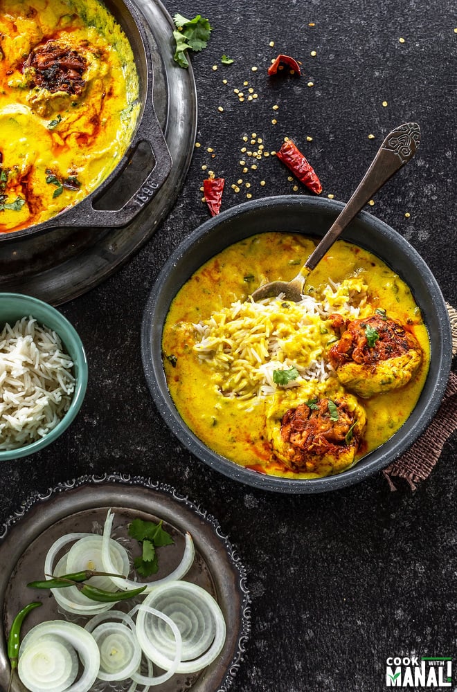 punjabi kadhi pakora served with rice in a black bowl along with a spoon with another plate of sliced onion and bowl of rice kept on the side