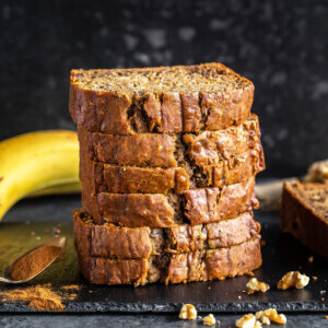 stack of banana bread slices placed on a black board with pieces of walnuts sprinkled around and a banana kept in the background