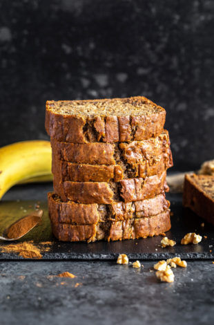stack of banana bread slices placed on a black board with pieces of walnuts sprinkled around and a banana kept in the background