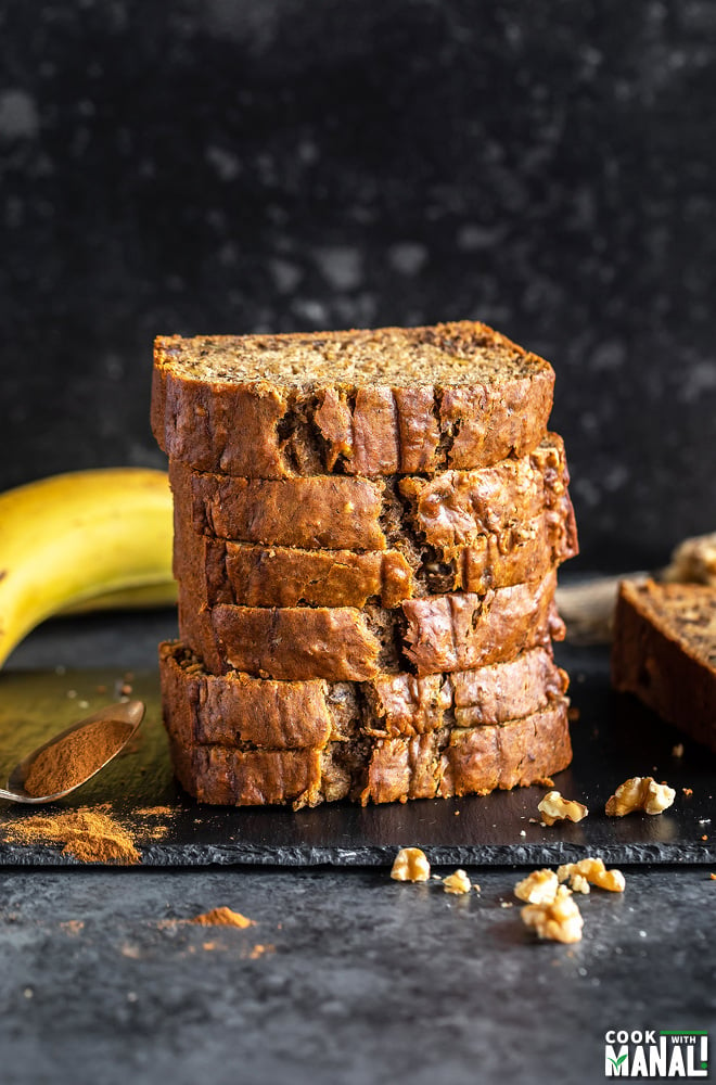 stack of banana bread slices placed on a black board with pieces of walnuts sprinkled around and a banana kept in the background